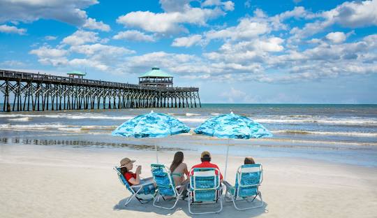 A group of tourists sitting by the edge of Folly Beach under beach umbrellas.
